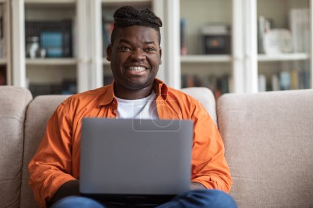 Photo for Positive overweight millennial african american man with braids in casual outfit sitting on sofa, typing on laptop keyboard, looking at copy space and smiling, male freelancer working from home - Royalty Free Image