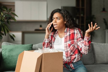 Photo for Shocked angry upset millennial black lady call by phone, arguing with delivery service in living room interior with box. Unpacking purchase, broken buy, mistake, problems with online shopping at home - Royalty Free Image