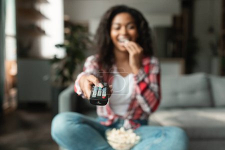 Photo for Happy millennial black lady with remote control change channel on tv, watch movie, eat popcorn, enjoy free time, sit on sofa in living room interior, blurred. Entertainment, fun at home, film evening - Royalty Free Image