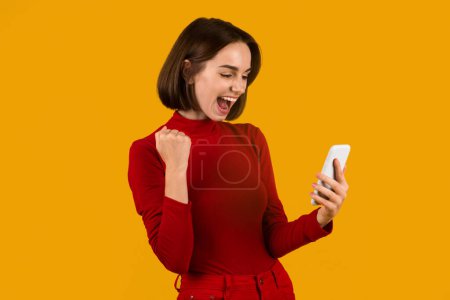 Foto de Mujer joven bonita emocional feliz en traje rojo casual celebrando el éxito sobre fondo de estudio naranja, sosteniendo el teléfono celular blanco, gritando y haciendo gestos, espacio para copiar. Apuesta en línea, apuestas, lotería - Imagen libre de derechos