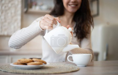 Photo for Happy young woman pouring herbal tea in mug, sitting at table in kitchen interior, selective focus. Food blog, housewife rest at home, visit and healthy breakfast during covid-19 outbreak - Royalty Free Image