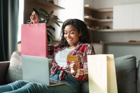 Photo for Cheerful millennial black female with many bags with purchases uses computer and credit card, enjoy delivery, sit on sofa in living room interior. Online shopping, big sale, cashback and finance home - Royalty Free Image