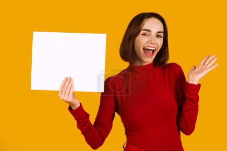 Foto de Feliz atractiva joven morena carismática en traje rojo que muestra el tablero de publicidad blanco en blanco con maqueta, muecas y gestos sobre fondo de estudio naranja, espacio de copia - Imagen libre de derechos