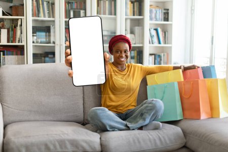 Foto de Feliz mujer negra bastante joven en rojo turbante shopaholic sentado en el sofá con bolsas de compras de colores, mostrando el teléfono con pantalla blanca en blanco en casa. Mockup para el concepto de compras en línea, espacio libre - Imagen libre de derechos