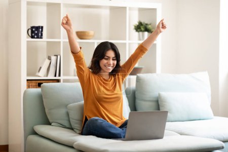 Foto de Buenas noticias. Mujer árabe joven feliz celebrando el éxito con el ordenador portátil mientras se sienta en el sofá en casa, Alegre a las mujeres de Oriente Medio levantando los puños y exclamando con emoción, Copiar el espacio - Imagen libre de derechos