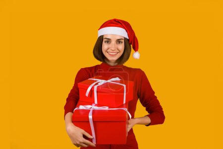 Foto de Feliz emocionado hermosa mujer joven en sombrero de Santa Claus con lindo peinado sosteniendo cajas de regalo rojas sobre fondo de estudio naranja, señora preparándose para la celebración de las fiestas de Navidad y Año Nuevo 2023 - Imagen libre de derechos