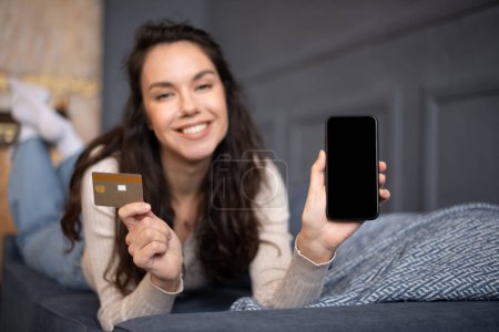 Téléchargez les photos : Heureuse femme caucasienne montrant smartphone avec écran vierge et carte de crédit, couchée sur le canapé dans l'intérieur du salon, maquette. Tech, achats en ligne et vente pendant covid-19 - en image libre de droit