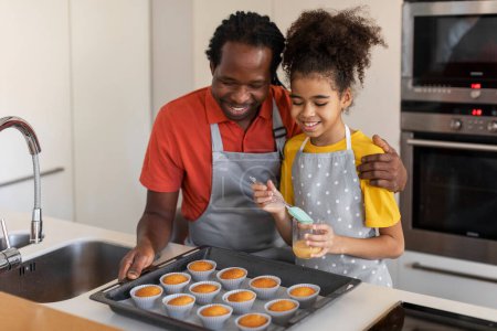 Téléchargez les photos : Noir papa et femelle enfant cuisson muffins ensemble à la maison, mignon preteen fille brossage haut de pâtisserie avec oeuf jaunes avant de le mettre au four, profiter de faire de la pâtisserie à la maison, gros plan - en image libre de droit