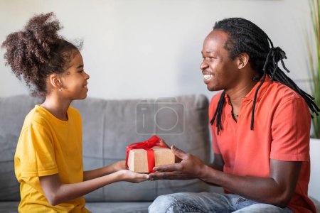 Foto de Día del Padre. Hija preadolescente negra dando caja de regalo a su padre en casa, joven afroamericano feliz recibiendo regalo de su hijo femenino mientras se sientan juntos en el sofá en la sala de estar - Imagen libre de derechos