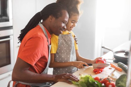 Photo for Cute Preteen Black Girl Making Vegetable Salad While Cooking With Father In Kitchen, Happy African American Family Dad And Daughter Preparing Healthy Food Together At Home, Side View Shot - Royalty Free Image