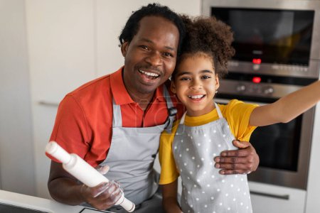 Foto de Linda chica negra tomando selfie con su papá mientras cocina en la cocina, feliz alegre padre afroamericano y preadolescente hija en delantales posando en la cámara mientras hornea en casa, primer plano - Imagen libre de derechos