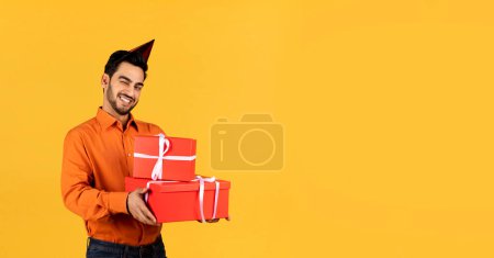 Foto de Feliz joven árabe sosteniendo cajas de regalo y guiños en la cámara, alegre guapo Oriente Medio masculino usando sombrero de fiesta de cumpleaños posando aislado sobre fondo amarillo, Panorama con espacio de copia - Imagen libre de derechos