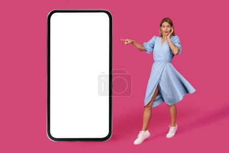 Photo for Shocked adult caucasian lady blonde in dress point finger at big smartphone with blank screen, isolated on pink background, studio. Online surprise, great news, sale, online app for blog, ad and offer - Royalty Free Image