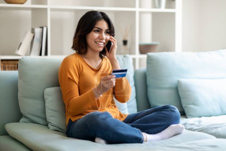 Foto de Concepto de comercio electrónico. Mujer árabe joven feliz sosteniendo la tarjeta de crédito y hablando en el teléfono celular mientras está sentado en el sofá en casa, sonriendo Oriente Medio Hembra haciendo compra en línea, Copiar el espacio - Imagen libre de derechos