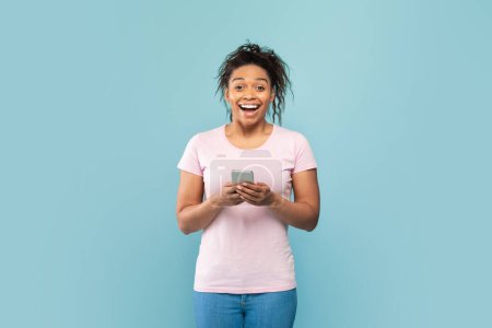 Téléchargez les photos : Surpris dame afro-américaine tenant smartphone et regardant la caméra dans l'excitation, loterie gagnante ou pari de casino en ligne sur fond de studio bleu. heureuse noir femme lecture incroyable nouvelles - en image libre de droit