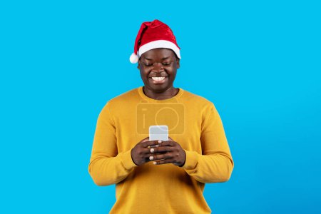 Photo for Happy Black Man Wearing Santa Hat Using Smartphone While Standing Isolated Over Blue Studio Background, Cheerful Young African American Guy Messaging Or Shopping Online Via Mobile Phone, Copy Space - Royalty Free Image