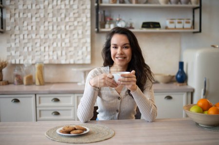 Téléchargez les photos : Portrait de femme caucasienne heureuse assise à la table de la cuisine avec une tasse de tisane et des biscuits, souriant à la caméra tout en appréciant la boisson chaude du matin à la maison, espace de copie - en image libre de droit