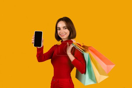 Photo for Cute attractive young woman with nice hairstyle in red outfit shopaholic purchasing online, holding colorful shopping bags and showing phone with black empty screen on orange background. E-commerce - Royalty Free Image