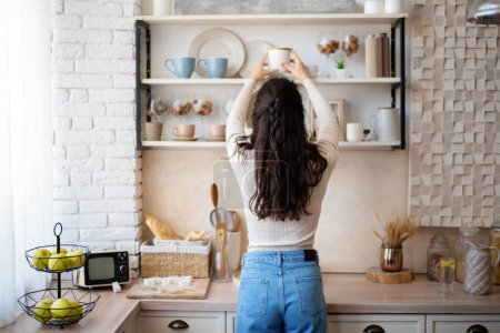 Téléchargez les photos : Jeune femme caucasienne mettre des assiettes et des bocaux sur l'étagère tout en se tenant dans l'intérieur de la cuisine domestique, vue arrière, espace libre. Concept de ménage - en image libre de droit