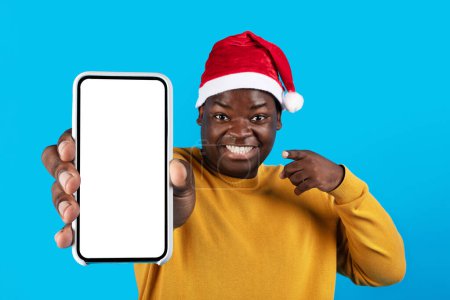 Téléchargez les photos : Annonce de Noël. Homme noir joyeux dans Santa Hat pointant vers Smartphone vide avec écran blanc à la main, Happy African American Guy Publicité Offre en ligne ou application mobile, Debout sur fond bleu, Mockup - en image libre de droit