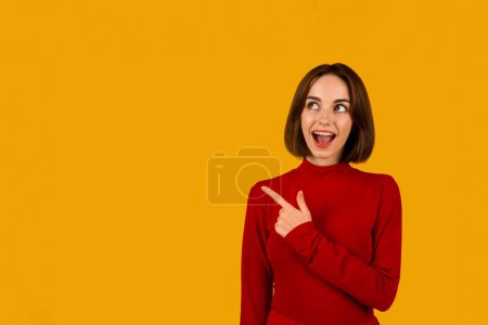 Foto de Cool excitado joven mujer caucásica con lindo peinado en rojo apuntando al espacio de copia para publicidad y muecas sobre fondo de estudio naranja, web-banner. Bonita oferta, descuento, noticias - Imagen libre de derechos