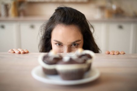 Foto de Mujer joven escondida detrás de la mesa y mirando deliciosos cupcakes en el plato, se centran en la señora. Concepto de apetito, dieta y gula - Imagen libre de derechos