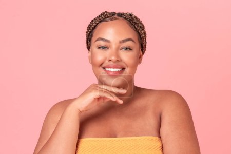 Foto de Concepto de belleza natural. Retrato de señora negra feliz más tamaño posando sobre fondo de estudio rosa y sonriendo a la cámara. Emocionada mujer afroamericana tocando la barbilla, disfrutando de una piel suave - Imagen libre de derechos