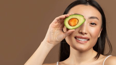 Photo for Portrait of young asian woman holding avocado hald and covering her eye with it, smiling at camera isolated on brown studio background, panorama with copy space - Royalty Free Image
