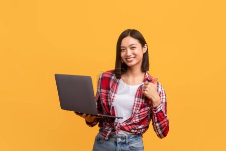 Téléchargez les photos : Femme asiatique excitée à l'aide d'un ordinateur portable et geste pouces levés, approuver le service en ligne, posant sur fond de studio jaune. J'aime ce concept de site web - en image libre de droit