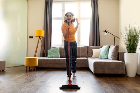 Photo for Positive Middle Eastern Female Wearing Wireless Headphones Having Fun While Cleaning Home, Cheerful Young Arab Woman Holding Broom And Using Handle As Microphone, Enjoying Domestic Chores, Copy Space - Royalty Free Image