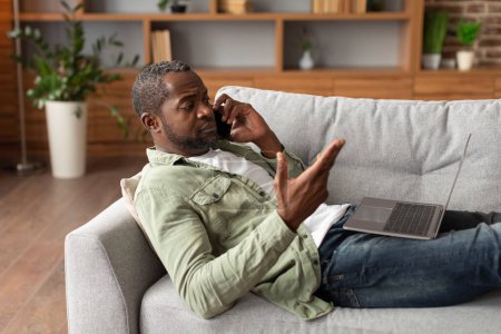 Photo for Serious unhappy middle aged black male calls by phone on sofa, gesticulate, watch at computer in living room interior. Problems with service, complaint, mistake in work remotely at home due covid-19 - Royalty Free Image