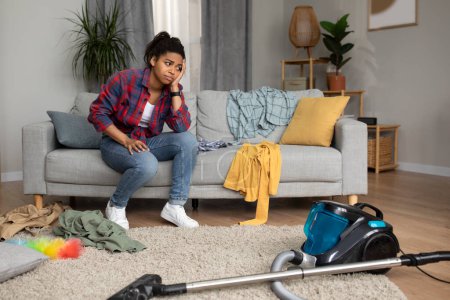 Photo for Tired sad millennial african american lady sit on sofa, look at vacuum cleaner and suffering from chaos and mess in living room interior. Household chores, problems with cleaning and many work at home - Royalty Free Image
