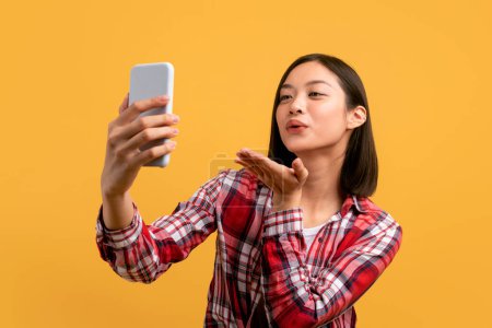 Foto de Alegre asiática dama tomando selfie con el teléfono celular, el envío de aire beso y la filmación de contenido para el blog en amarillo fondo del estudio. Coqueta mujer charlando en línea con el novio - Imagen libre de derechos