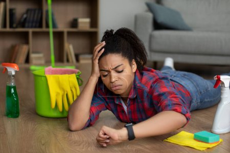 Photo for Sad tired millennial african american female lies on floor with cleaning supplies, looks at watch in room interior. Mess, waiting service for cleaning and household chores, late, a lot of work at home - Royalty Free Image
