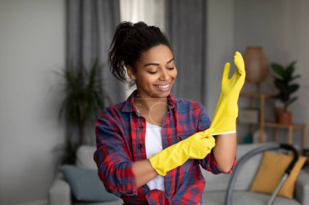 Téléchargez les photos : Joyeux millénaire dame afro-américaine met des gants en caoutchouc et prêt pour les tâches ménagères dans le salon intérieur. Service de nettoyage professionnel, hygiène, style de vie et beaucoup de travail à la maison - en image libre de droit