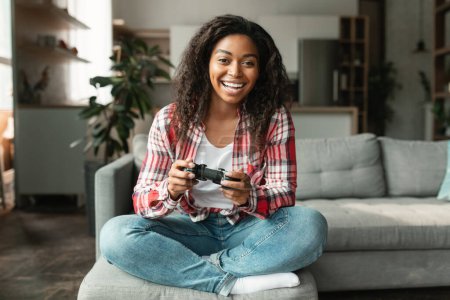 Photo for Glad pretty millennial black lady with joystick playing computer games, sitting on sofa alone, enjoy free time in living room interior, copy space. Entertainment and fun at home with modern technology - Royalty Free Image