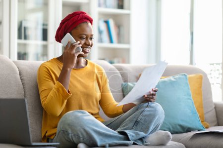 Téléchargez les photos : Femme d'affaires afro-américaine jeune femme attrayante avec turban rouge sur la tête en tenue décontractée assis sur le canapé avec ordinateur portable, lire les papiers, travailler à la maison, avoir une conversation téléphonique, copier l'espace - en image libre de droit