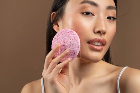 Photo for Face cleansing concept. Closeup of korean lady using soft facial cellulose sponge, washing face isolated on brown studio background, free space - Royalty Free Image