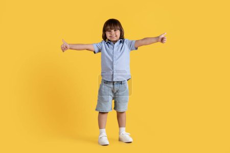 Téléchargez les photos : Bonne idée. Adorable petit garçon excité montrant pouce en l'air, étendant largement ses mains et souriant, fond de studio orange, pleine longueur, espace vide - en image libre de droit