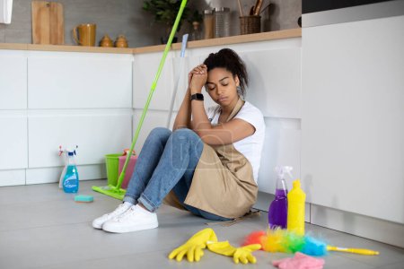Téléchargez les photos : Fatiguée millénariste afro-américaine malheureuse dans le tablier s'assoit sur le sol avec des fournitures de nettoyage, dort les yeux fermés dans l'intérieur de la cuisine minimaliste. Chaos, sale et problèmes avec le nettoyage de la maison - en image libre de droit
