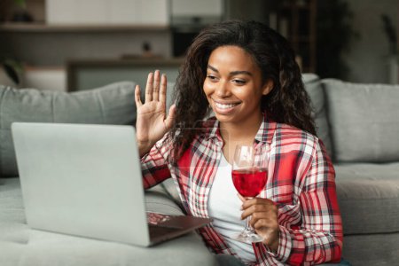 Glad millennial black lady waving hand, say hello greeting in computer camera, hold glass of wine in living room interior. Holiday celebration, congratulation, date remote with technology at home