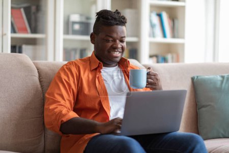 Photo for Domestic entertainment concept. Relaxed positive young black guy in casual sitting on couch with computer on lap, holding mug, watching movie or websurfing and drinking tea at home, copy space - Royalty Free Image