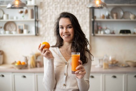 Photo for Happy woman drinking orange juice and holding fruit, standing in kitchen interior, free space. Young lady drinking juice at home. Fit smiling female preparing healthy fruit juice - Royalty Free Image