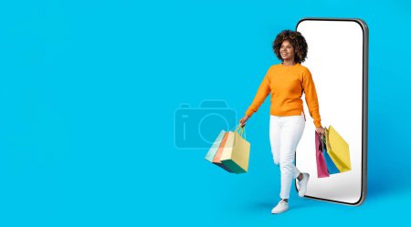Photo for E-commerce. Happy cheerful young black woman in casual with bushy hair shopaholic walking out of big phone with white empty blank screen, smiling, carrying shopping bags, blue background, copy space - Royalty Free Image