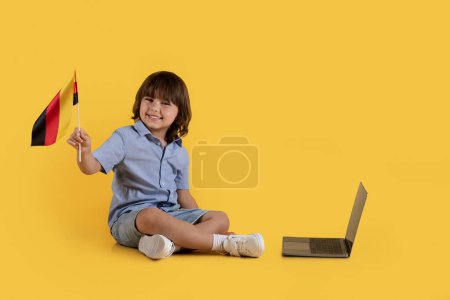 Foto de Educación en línea en el extranjero. Divertido niño feliz cuidado bandera de Alemania y sonriendo a la cámara, sentado cerca de la computadora portátil con clases de aprendizaje, fondo de estudio naranja, espacio libre - Imagen libre de derechos