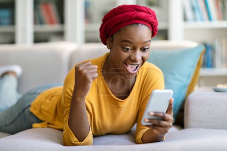 Téléchargez les photos : Émotionnel heureux afro-américain jeune femme portant turban rouge traditionnel à l'aide d'un smartphone à la maison, couché sur le canapé dans le salon confortable, en regardant l'écran du gadget et des gestes, de bonnes nouvelles, espace libre - en image libre de droit
