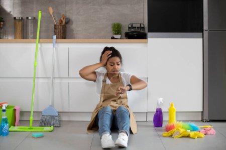 Photo for Tired sad surprised millennial african american lady sitting on floor with cleaning supplies, looking at watch in minimalist kitchen interior. Late with cleaning, lifestyle, chaos and disorder at home - Royalty Free Image