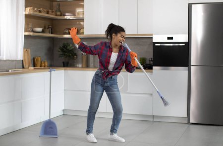 Photo for Happy millennial african african lady superstar in rubber gloves sings in mop, imaginary microphone, mopping floor, enjoy household chores in kitchen interior. House cleaning alone at weekend at home - Royalty Free Image