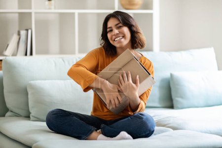 Photo for Delivery Concept. Happy Arab Man Holding Big Cardboard Box At Home, Cheerful Young Middle Eastern Female Embracing Delivered Parcel And Smiling At Camera While Sitting On Couch In Living Room - Royalty Free Image