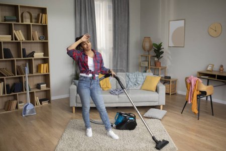 Photo for Tired sad millennial african american housewife wipes forehead and with vacuum cleaner, suffers from household chores in living room interior. A lot of work at home, hygiene and problems with cleaning - Royalty Free Image